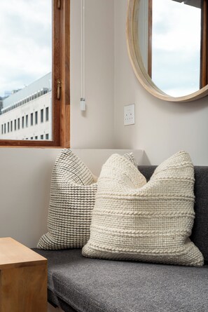 A room with a grey sofa, two white knitted pillows, a round mirror, and a window with a view of buildings. - Charlie and the Chairman | Contemporary Suite (Cape Town)