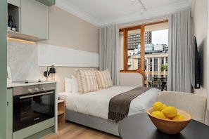 A modern hotel room with a bed, a wooden table with lemons, a built-in oven, and a view of the city. - Charlie and the Chairman | Designer Suite in CBD (Cape Town)