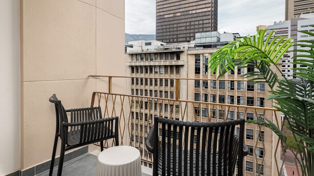 Ein Balkon mit zwei schwarzen Stühlen und einem kleinen Tisch, mit Blick auf eine Stadtlandschaft mit hohen Gebäuden und einer Palme.