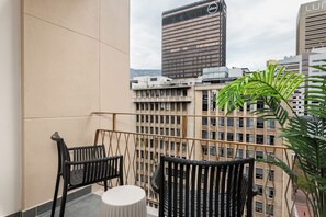 Ein Balkon mit zwei schwarzen Stühlen und einem kleinen Tisch, mit Blick auf eine Stadtlandschaft mit hohen Gebäuden und einer Palme.
