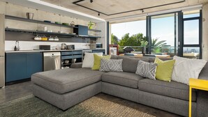 Een moderne keuken met een grijze hoekbank, een gele bijzettafel en een groot raam met uitzicht op de omgeving.