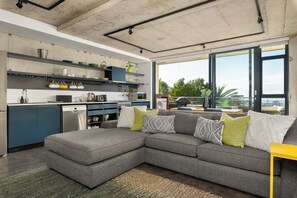 A modern kitchen with a grey sectional sofa, a yellow side table, and a large window offering a view of the outdoors. - Skyline Escape | Views, Pool and Braai (Cape Town)