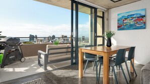 Een balkon met een barbecue, een eettafel met stoelen, een schilderij aan de muur en uitzicht op de stad.