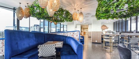 A modern lounge area with a blue sofa, hanging crystal chandeliers, and a green leafy installation.