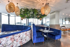 Ein modernes Restaurant mit blauen Samssitzbänken, trennenden Elementen mit Blumenmuster und hängenden Leuchten.