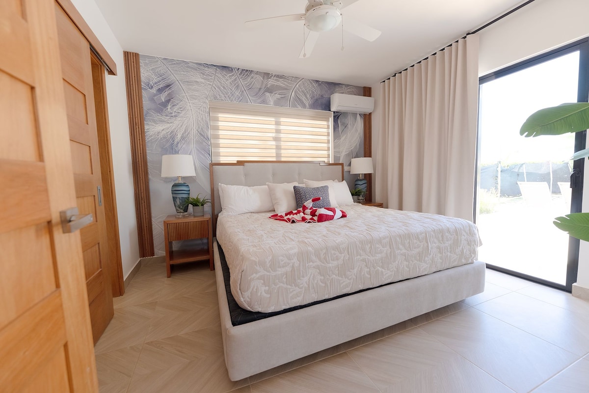 Ein modernes Schlafzimmer mit einem großen Bett, einem Deckenventilator, einem Fenster mit Vorhängen und einer wandmontierten Klimaanlage.