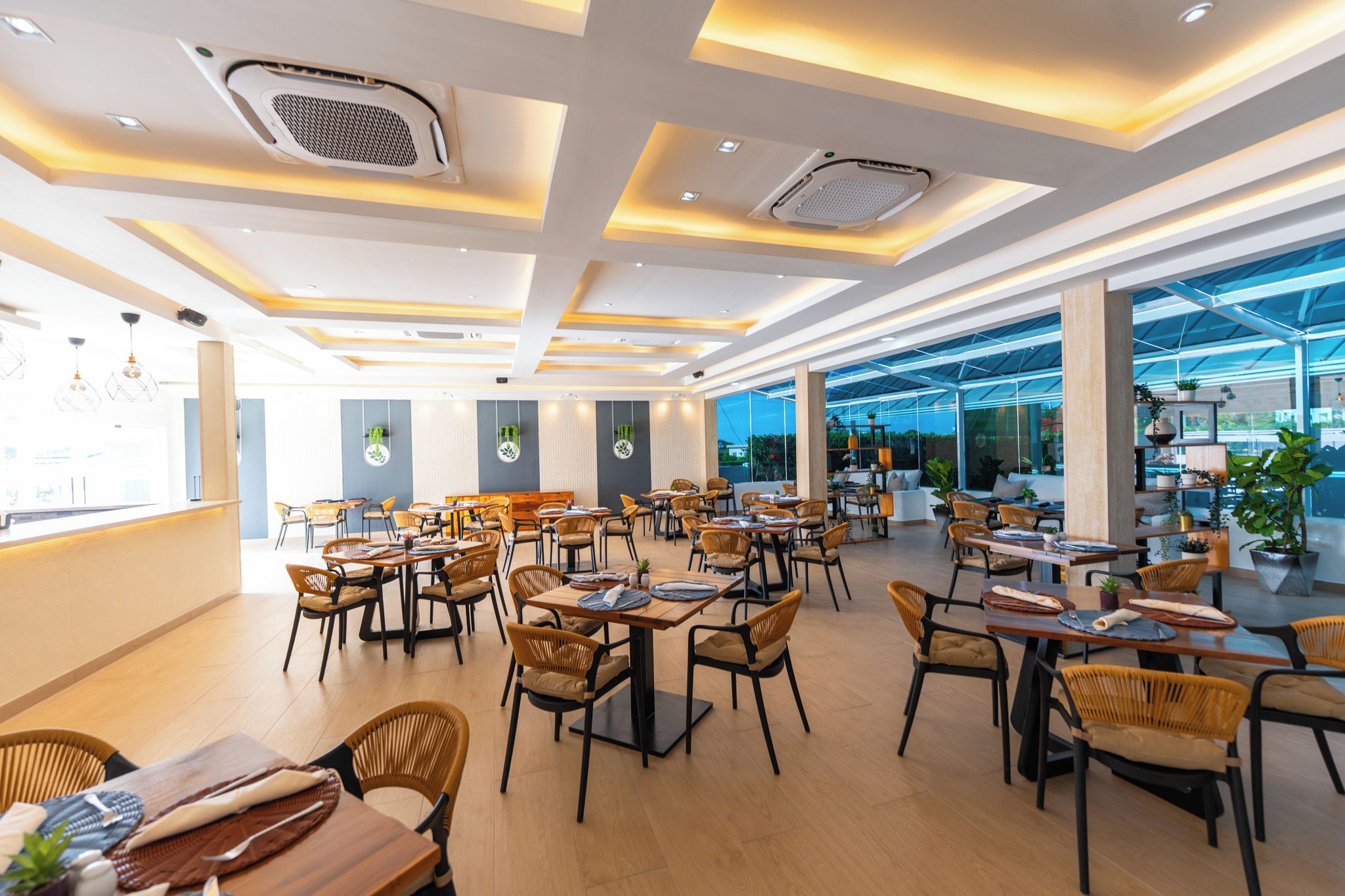 Ein modernes Restaurant-Interieur mit mehreren Tischen, Stühlen, Lüftungsschlitzen an der Decke und großen Fenstern.