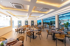 Ein modernes Restaurant-Interieur mit mehreren Tischen, Stühlen, Lüftungsschlitzen an der Decke und großen Fenstern.