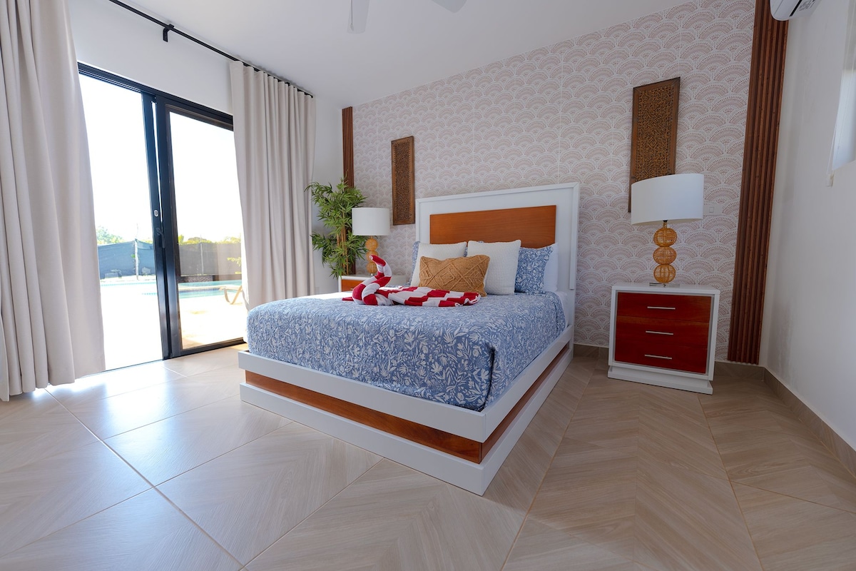 Ein modernes Schlafzimmer mit einem großen Bett, einem Nachttisch, einer Lampe und Blick auf einen Swimmingpool.
