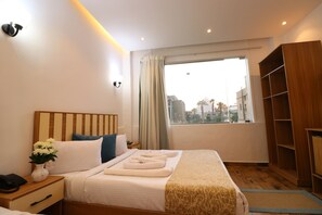 A hotel room with a bed, a nightstand, a bedside table, a window with curtains, and a wooden wardrobe.