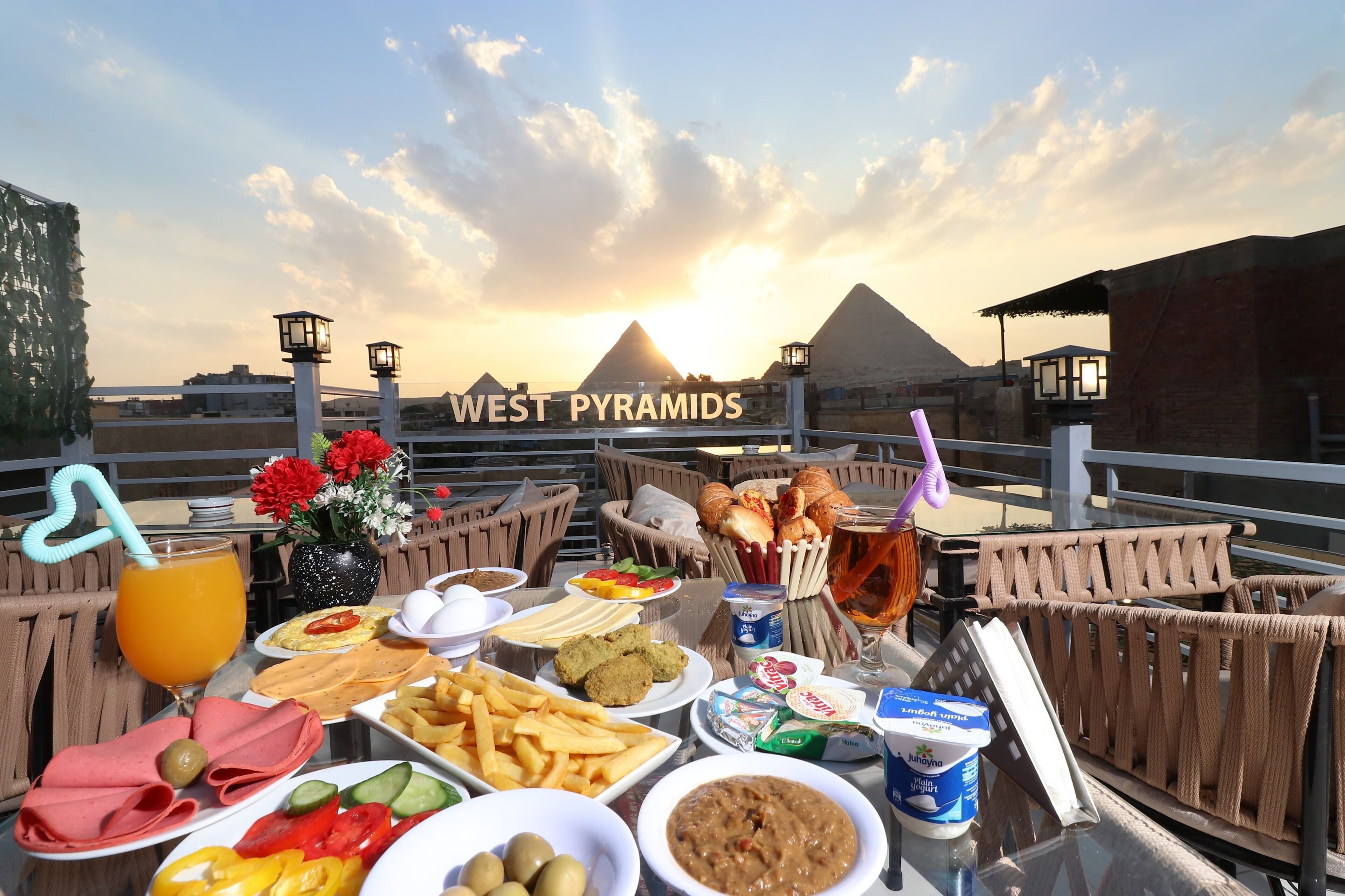 A dining table with food and drinks, a view of the West Pyramids, and a sunset.
