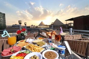 A dining table with food and drinks, a view of the West Pyramids, and a sunset.