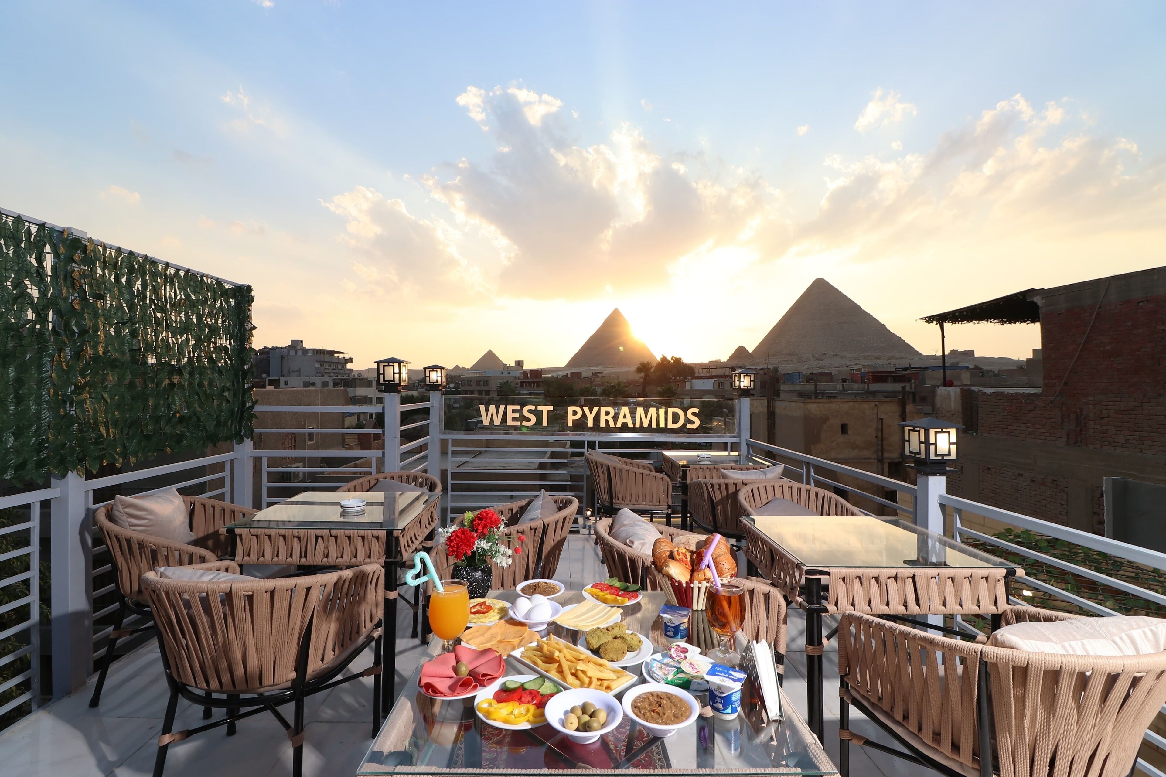 Un espacio de comedor en la azotea con muebles de mimbre, una mesa preparada con comida y bebidas, y las Pirámides de Giza al fondo.
