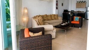 Ein Wohnzimmer mit einer Couch, einem Korbsessel, einem Couchtisch und einer Küchenzeile mit Schränken und Geräten.