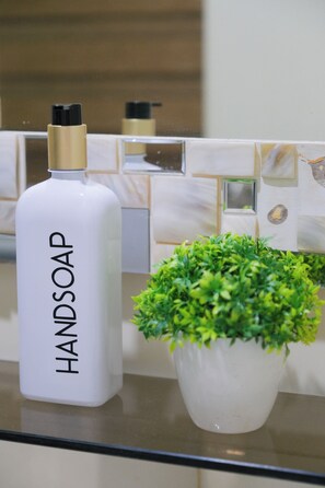 Une salle de bain avec un distributeur de savon blanc portant l’inscription « SAVON POUR LES MAINS » et une plante en pot posée sur le comptoir.