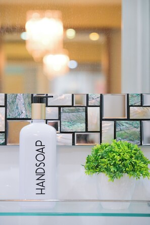 Ein Badezimmer mit einer weißen Flasche mit dem Aufdruck "HANDSOAP" auf einer Glastafel, einer kleinen Topfpflanze und einer gefliesten Spritzschutzleiste mit geometrischem Muster.