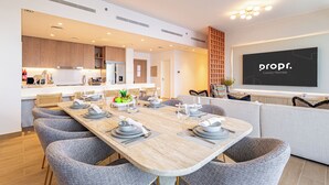 Une cuisine moderne avec des meubles en bois, un coin repas doté d’une longue table dressée pour six personnes, et un salon avec un grand écran diffusant des images de « propf. Luxury Homes ».