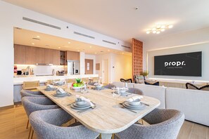 Eine moderne Küche mit Holzausstattung, ein Essbereich mit einem langen Tisch, gedeckt für sechs Personen, und ein Wohnbereich mit einem großen Bildschirm, der 'propf. Luxury Homes' zeigt.