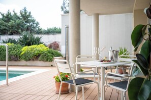 En udendørs terrasse med swimmingpool, hvide metalstole, et bord med en metalkande og plantekrukker.