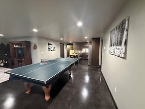 A room with a ping pong table, a kitchen area, and a dining table with chairs.