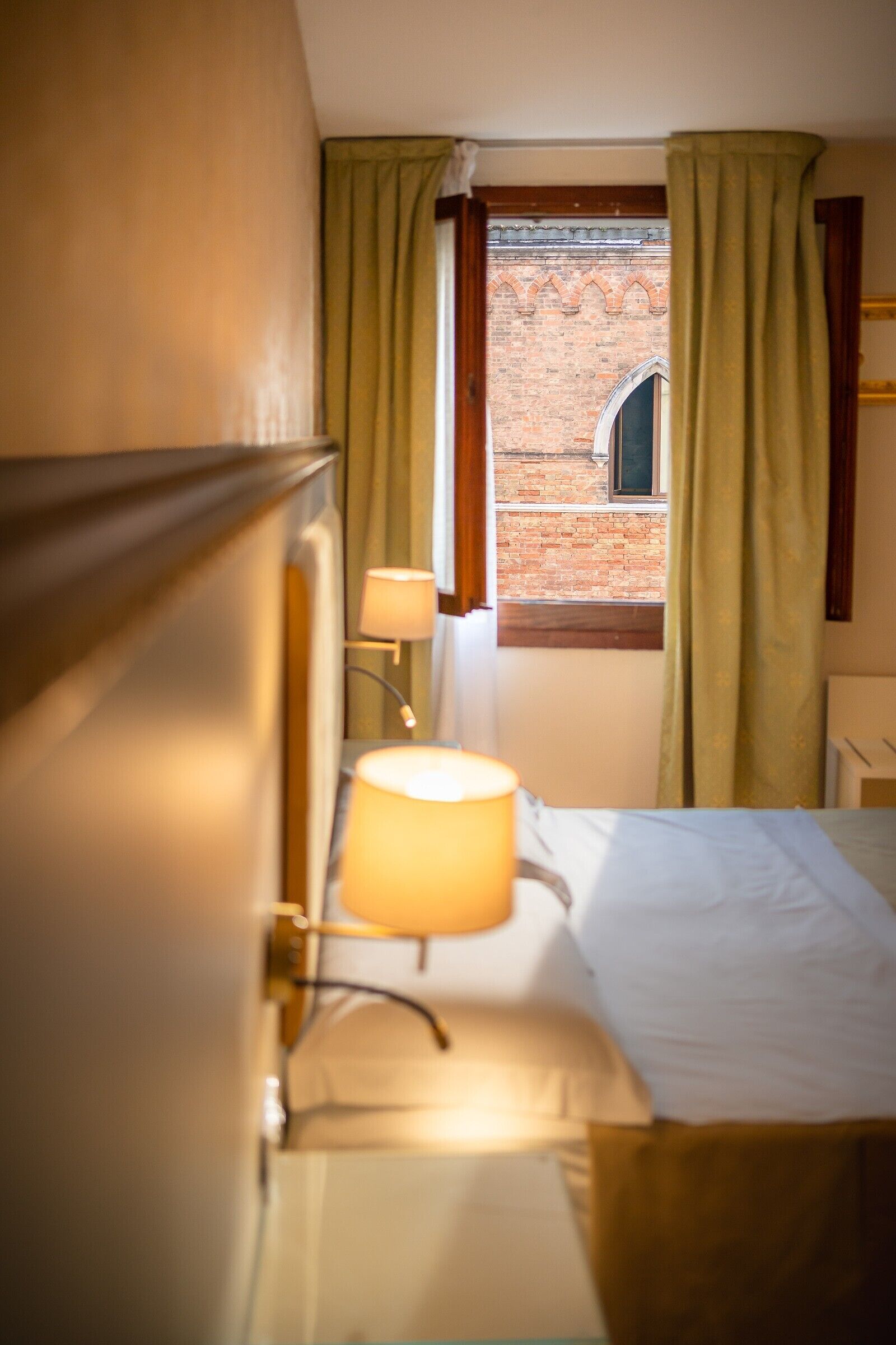 Ein Hotelzimmer mit Bett, Lampe, Fenster mit Vorhängen und Blick auf ein Backsteingebäude.