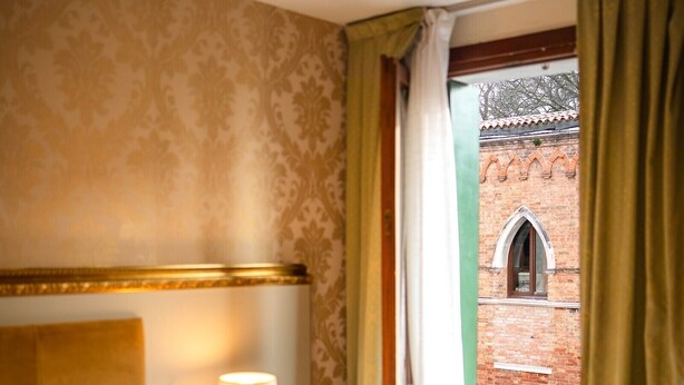 Ein Hotelzimmer mit Bett, Nachttisch mit Lampe, Fenster mit Vorhängen und Blick auf ein Backsteingebäude.