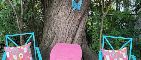 Two turquoise chairs with patterned cushions, a small pink bench, and two blue butterfly decorations on a large tree.
