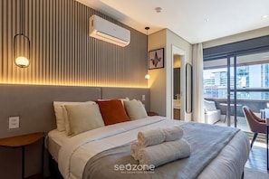 Ein Hotelzimmer mit Bett, Nachttischen, einer Klimaanlage, einer Wandleuchte und Blick auf die Stadt durch große Fenster.