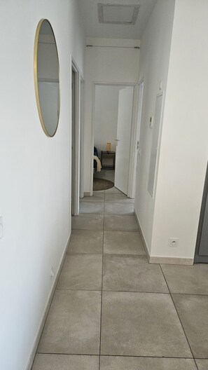 A corridor with a tiled floor, a round mirror on the left, and a door at the end leading to another room.