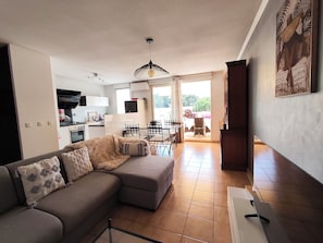 Ein Wohnzimmer mit einer Couch, ein Essbereich mit Stühlen und einem Tisch, eine Küche mit Geräten und ein Blick durch eine offene Tür.