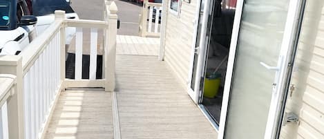 A wooden deck leading to a building with a sliding glass door, a white fence, and parked cars in the background.