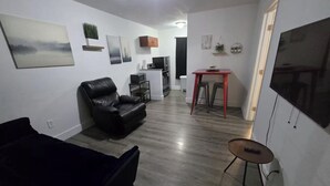Ein Wohnzimmer mit einer Couch, einem Sessel, einem kleinen Tisch und einem Fernseher. Im Hintergrund ist eine Küchenzeile zu sehen.