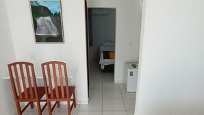 Una habitación con una cama, una mesita, dos sillas, un cuadro en la pared y una puerta que da a otra habitación.