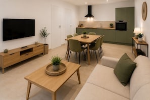 Een moderne woonkamer met een flatscreen televisie, houten meubels, een eethoek met stoelen, een keuken met fornuis en spoelbak, en een groene accentmuur.