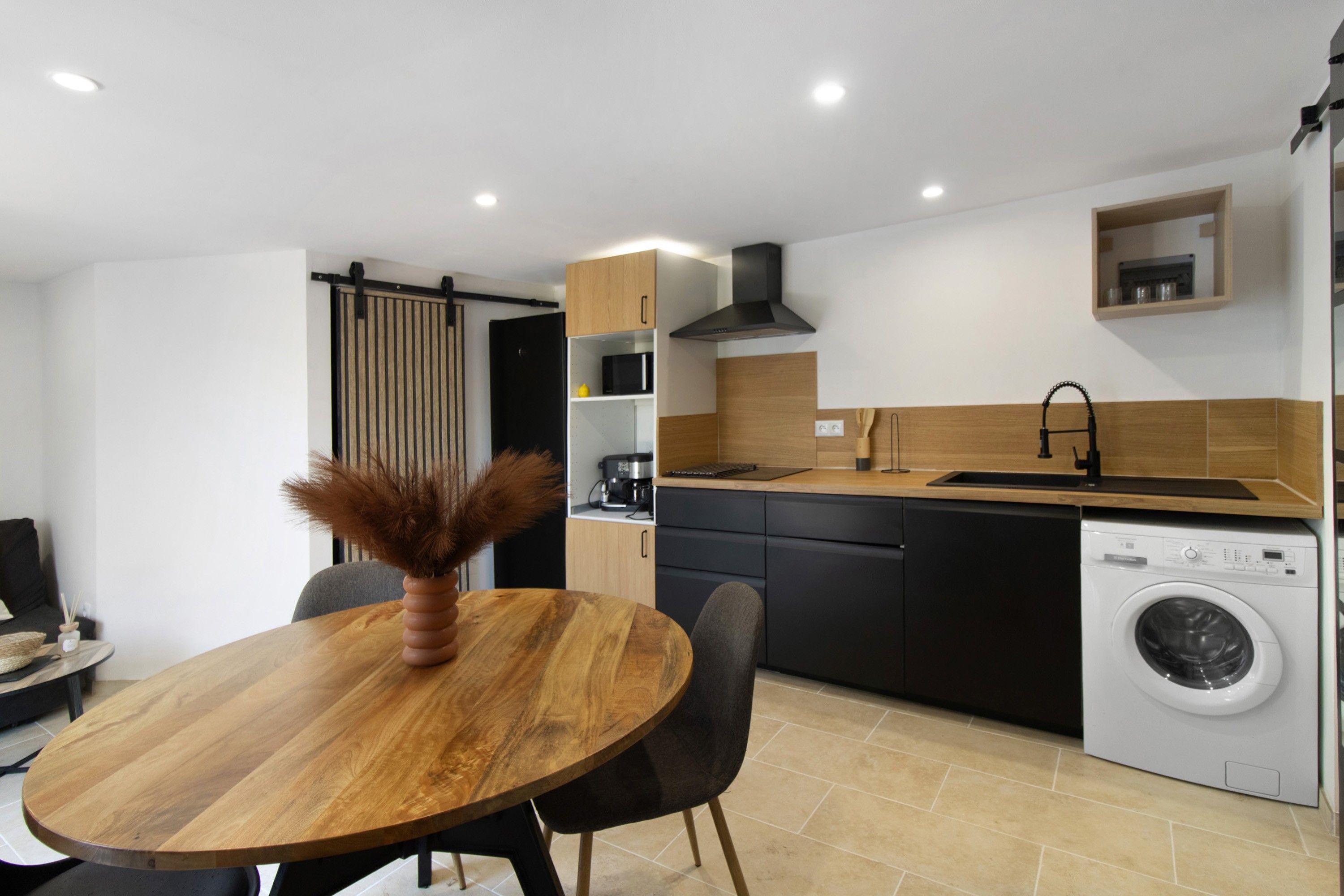 A modern kitchen with a wooden dining table, black chairs, a built-in oven, a microwave, and a washing machine.
