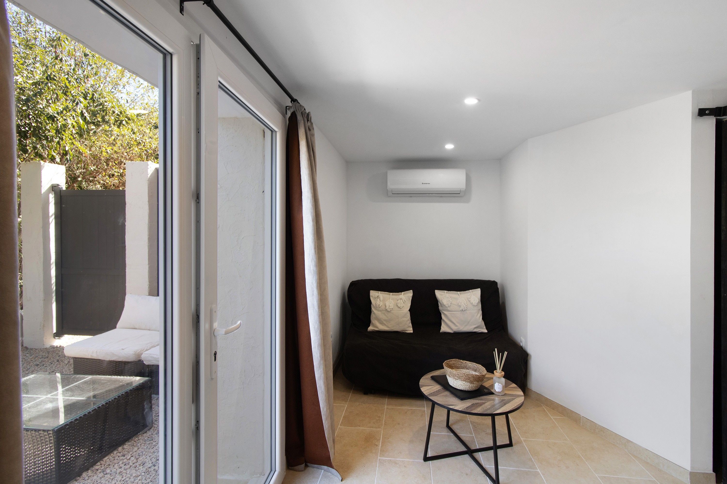 A modern living room with a glass door leading to an outdoor area, a black sofa with decorative pillows, and a small round table with a woven basket and reed diffuser.