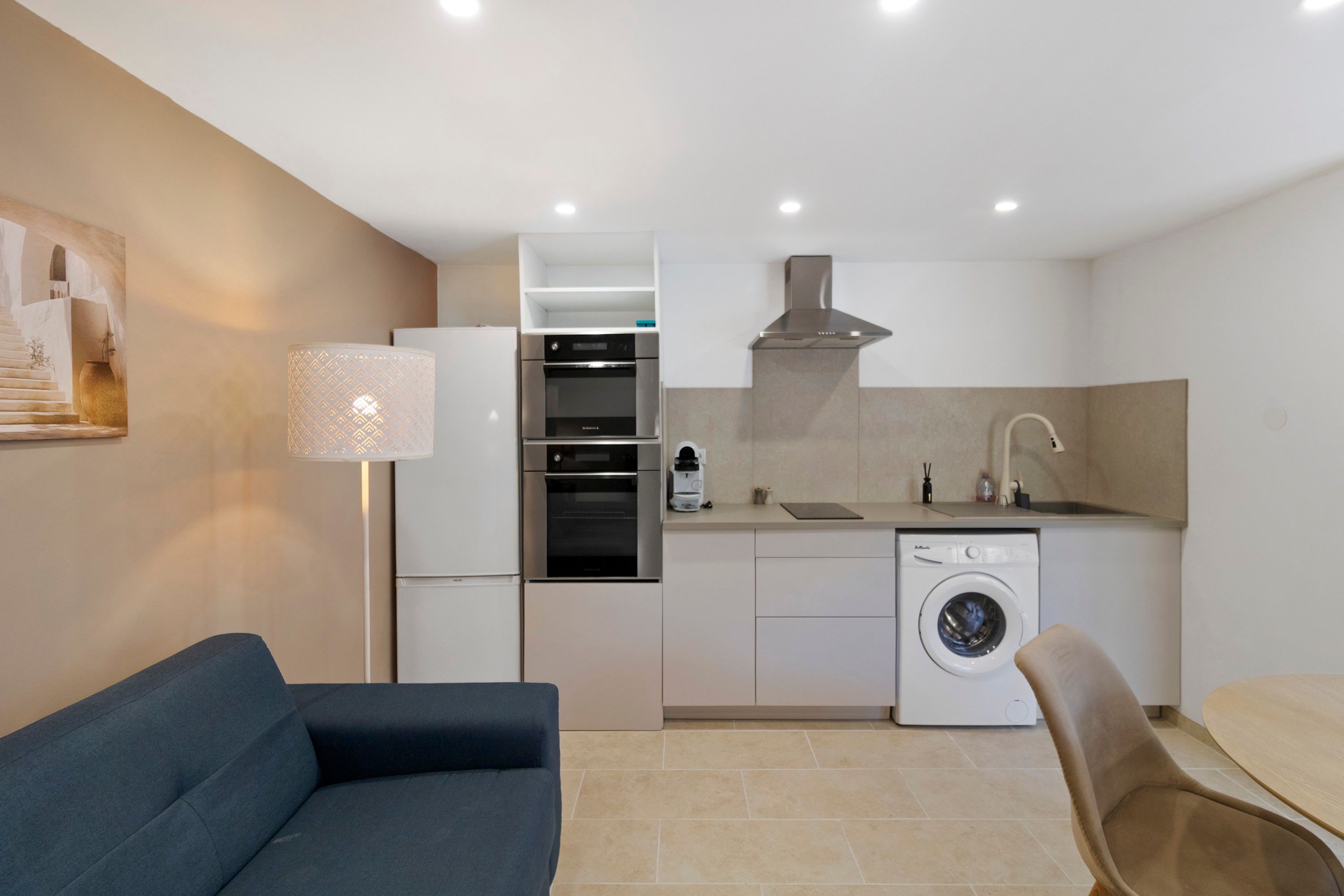 A compact kitchen with a washing machine, a coffee maker, and a dining area with a sofa and chair.