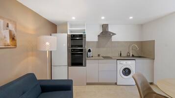A compact kitchen with a washing machine, a coffee maker, and a dining area with a sofa and chair.