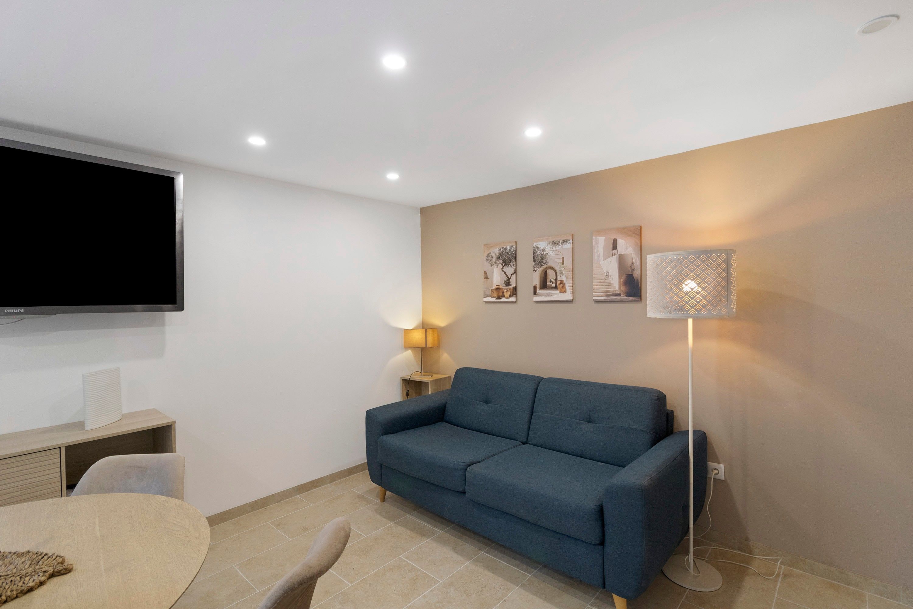 Sala de estar com sofá azul, TV de tela plana, abajur e três quadros na parede.