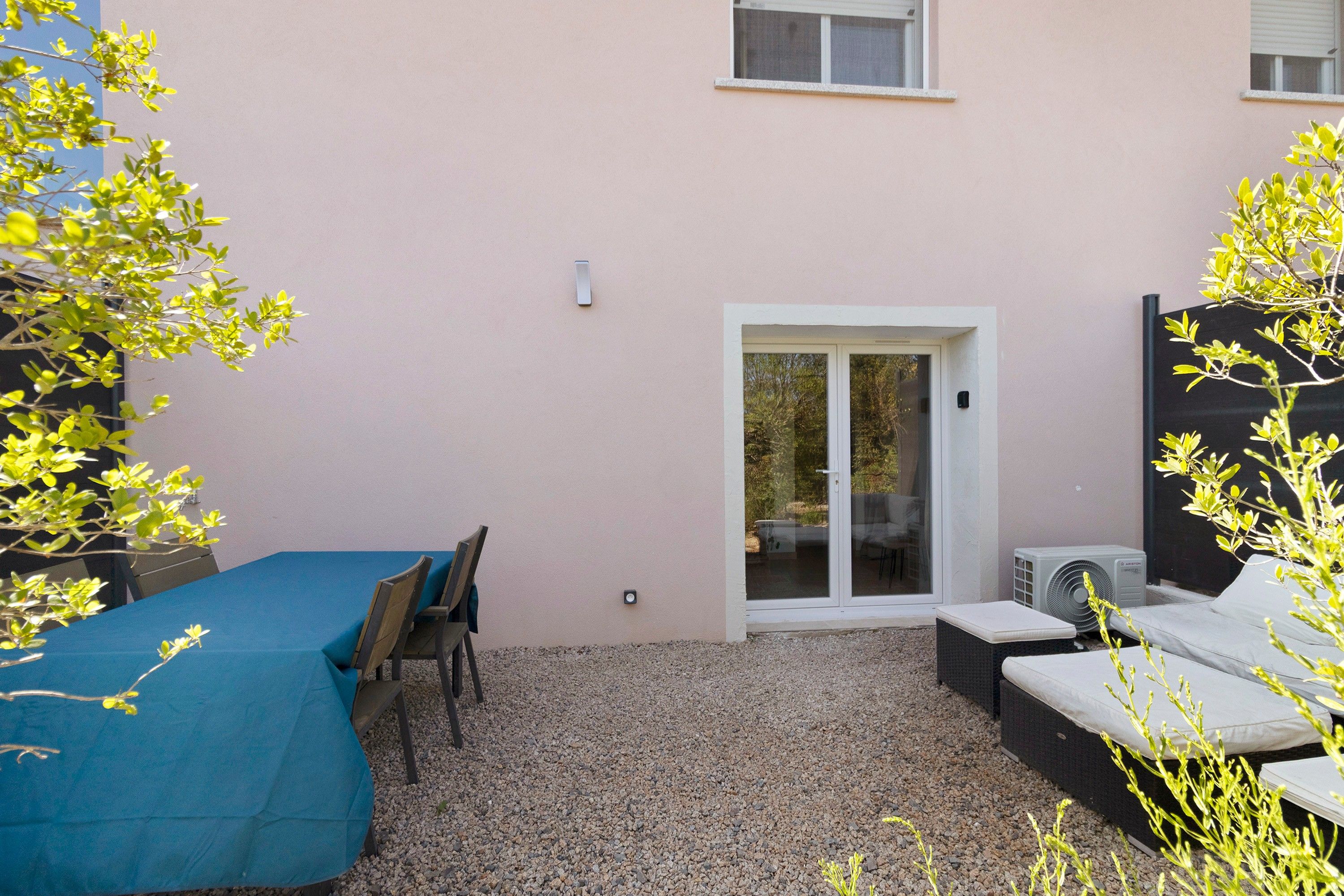 Un espace patio avec une table et des chaises, une porte-fenêtre coulissante donnant sur un espace extérieur abrité, et un petit climatiseur.