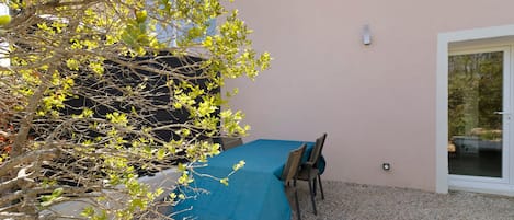 A covered outdoor seating area with a table and chairs, surrounded by gravel and a tree.