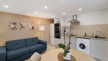 Uma sala de estar moderna com um sofá azul, uma mesa de jantar com uma planta e uma cozinha equipada com máquina de lavar e forno.