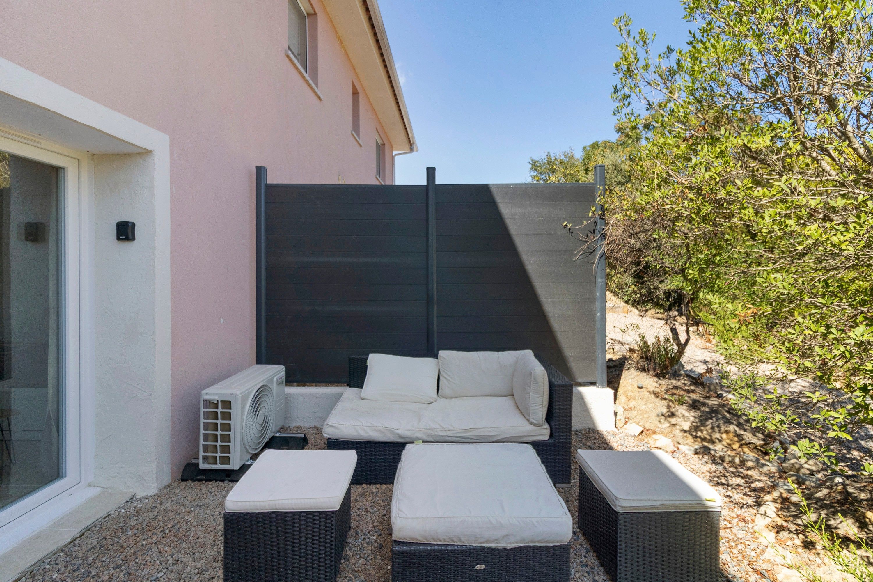 A private outdoor seating area with a sofa and two ottomans, a small air conditioning unit, and a glass door entrance.