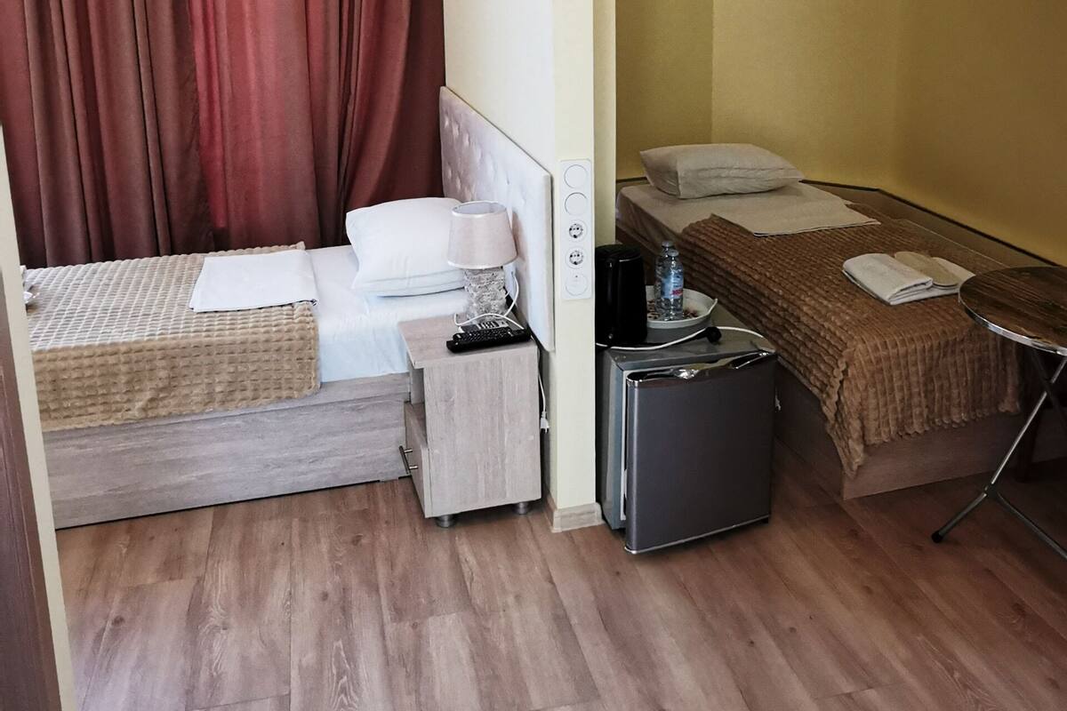 A compact hotel room with a bed, a small table, a refrigerator, and a bedside table with a lamp.