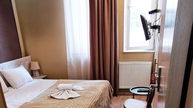 A hotel room with a bed, a chair, a small table, a lamp, a window with curtains, and a radiator. - Tbiliseli Inn (Tbilisi)