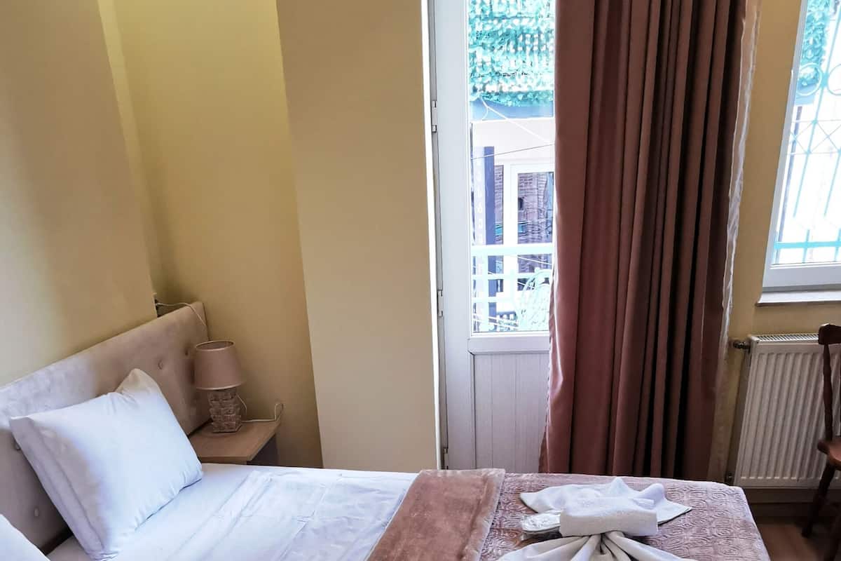 A hotel room with a bed, a nightstand, a window with curtains, and a view of buildings outside.