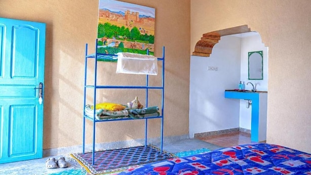 Ein Zimmer mit blauer Tür, einem Drahtgestell mit Handtüchern, einem Landschaftsbild, einem Waschbecken mit Spiegel und einem gemusterten Teppich.