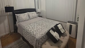 Une chambre à coucher comprenant un lit, une table de chevet, une lampe, un ventilateur de plafond, un radiateur et un banc.