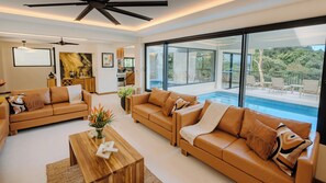 A modern living room with leather sofas, a wooden coffee table, and a large window overlooking a pool and outdoor seating area. - Luxury Brand New : Casa Rambutan - 3BR (Quepos)