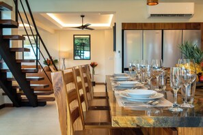 A dining area with a long table set for six, wooden chairs, and a modern kitchen in the background. - Luxury Brand New : Casa Rambutan - 3BR (Quepos)